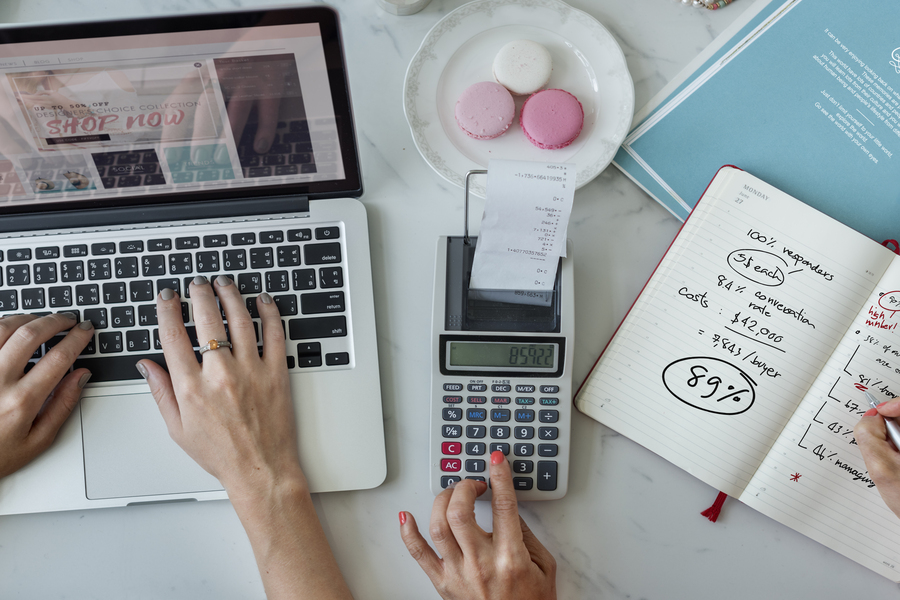 Two pairs of hands of Businesswomen calculating Esty fees on a laptop, calculator, and notebook.