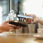 A customer's hand holding a smartphone with the Google Wallet and paying on a card terminal.